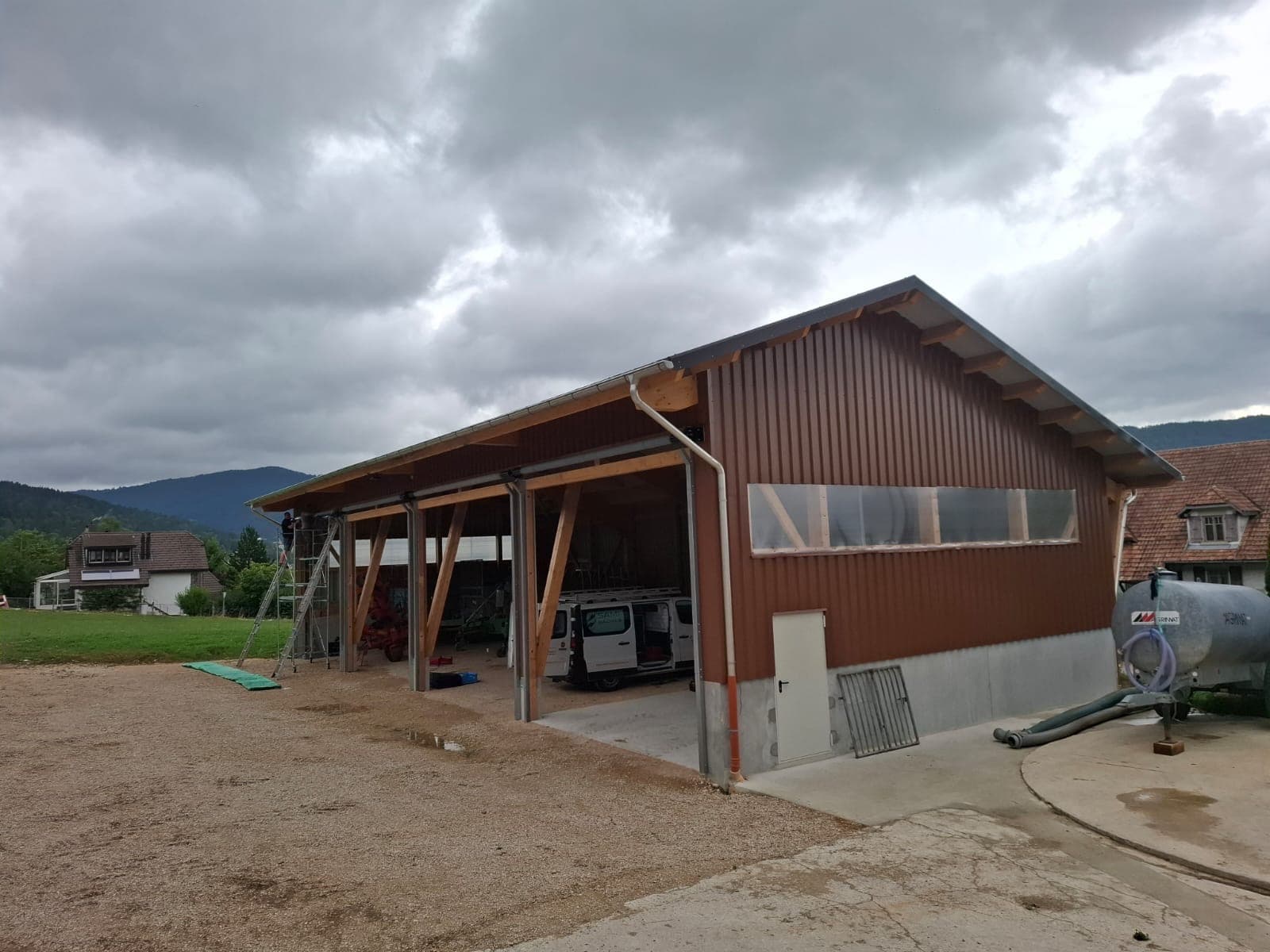 Completed agricultural hall with brown corrugated metal cladding, Swiss mountains in background